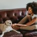Side view of young cheerful African American female conversing on cellphone while caressing dog on leather couch at home