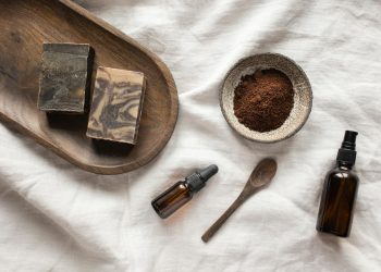 Top view of coffee scrub in bowl composed with bottles of aromatic oil on white sheet